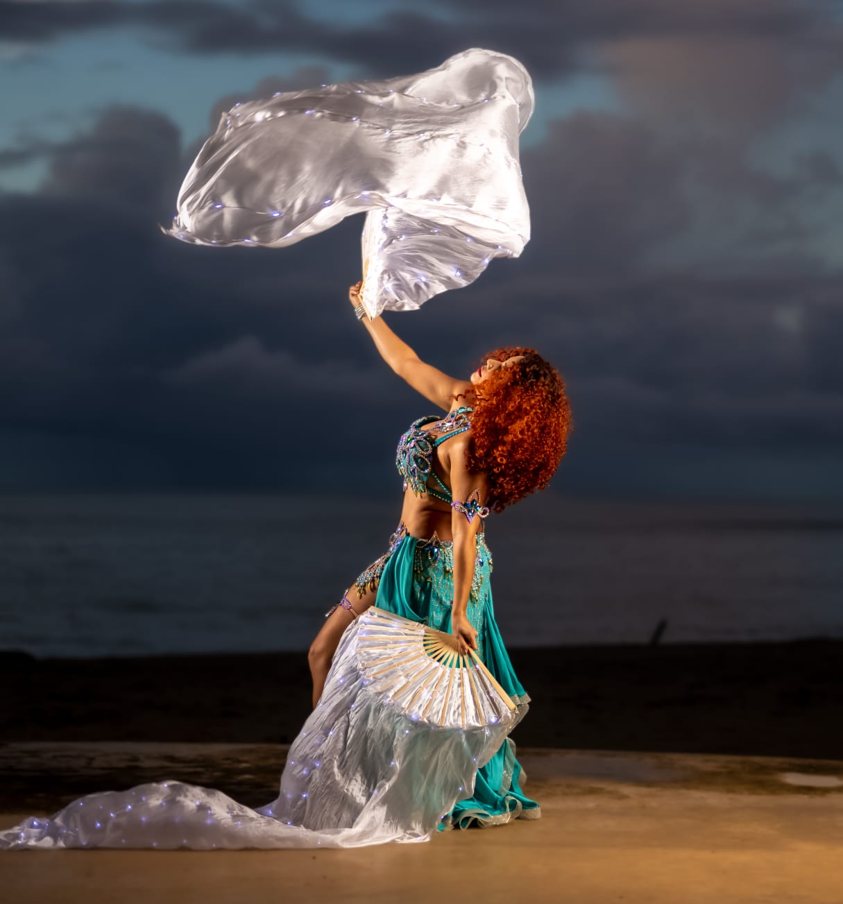 Adira Yamilet bailando belly dance con abanicos de velo plateados iluminados en una playa al atardecer.